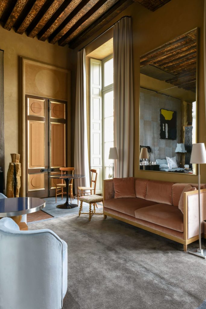 Salon élégant d'une suite de l'hôtel Cour des Vosgesavec canapé rose, miroir mural, table et chaises, rideaux beiges, poutres bois.