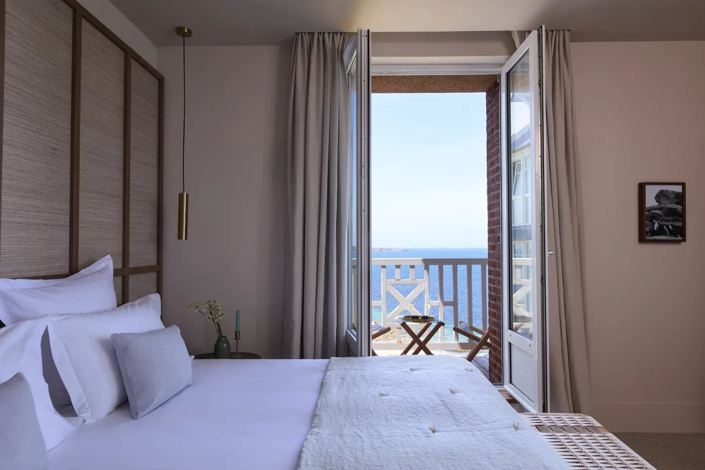 Une chambre de l'hôtel Les Bassans ouverte sur un balcon face à la mer, tête de lit en fibres naturelles et linge blanc.