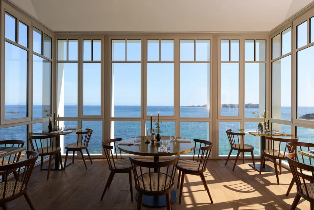 La salle à manger de l'hôtel Les Bassans, tables de laiton et chaises cannées face aux grandes baies ouvertes sur la baie de Perros-Guirec.