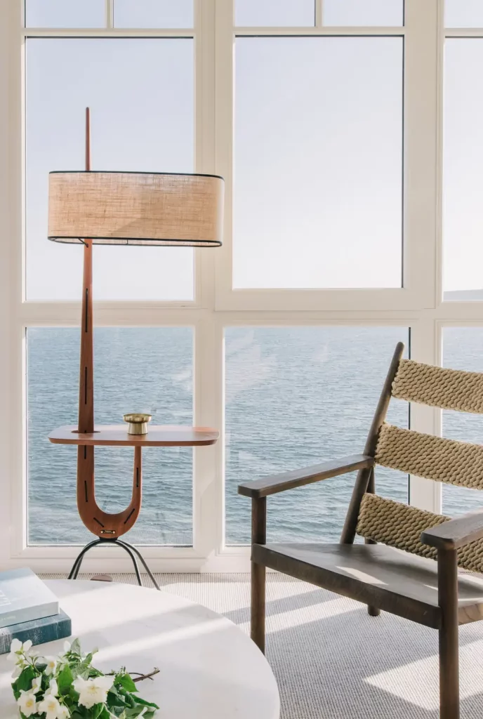 Un coin salon de l'hôtel Les Bassans, lampadaire vintage en teck à tablette et fauteuil de corde tressée, face aux baies ouvertes sur la mer.