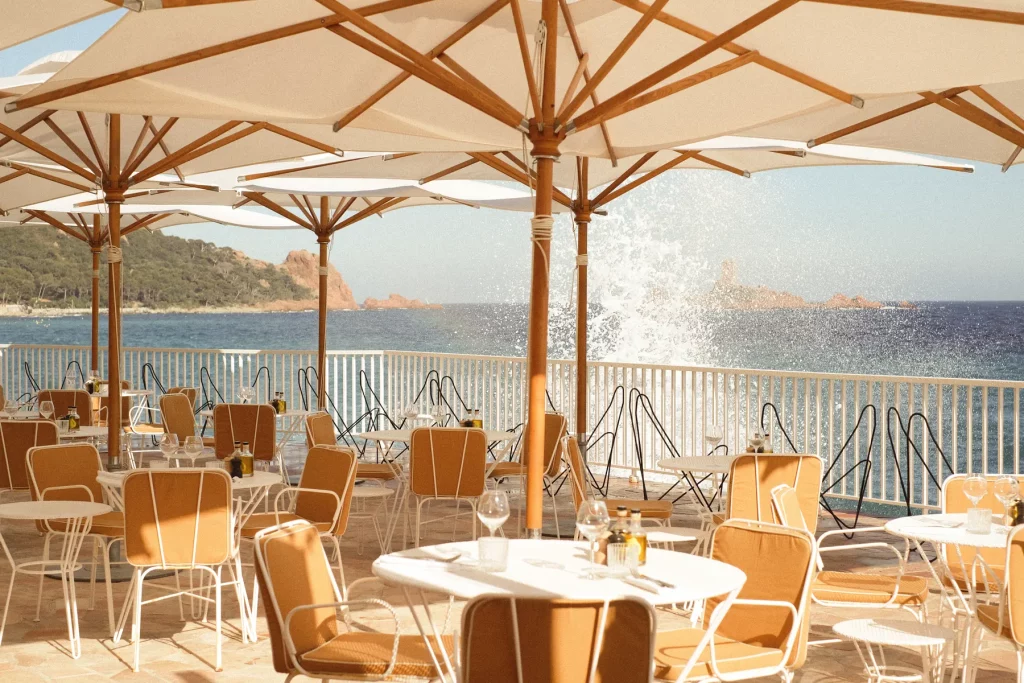 Terrasse du restaurant des Roches Rouges en bord de mer : tables rondes, chaises jaunes et parasols de toile écrue, une vague éclatant contre la digue.