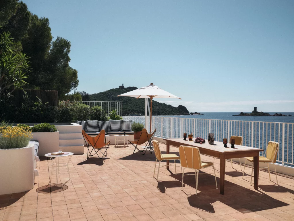 Grande table en bois et chaises colorées sur une terrasse en terre cuite des Roches Rouges, face aux roches de l'Estérel et à la mer.