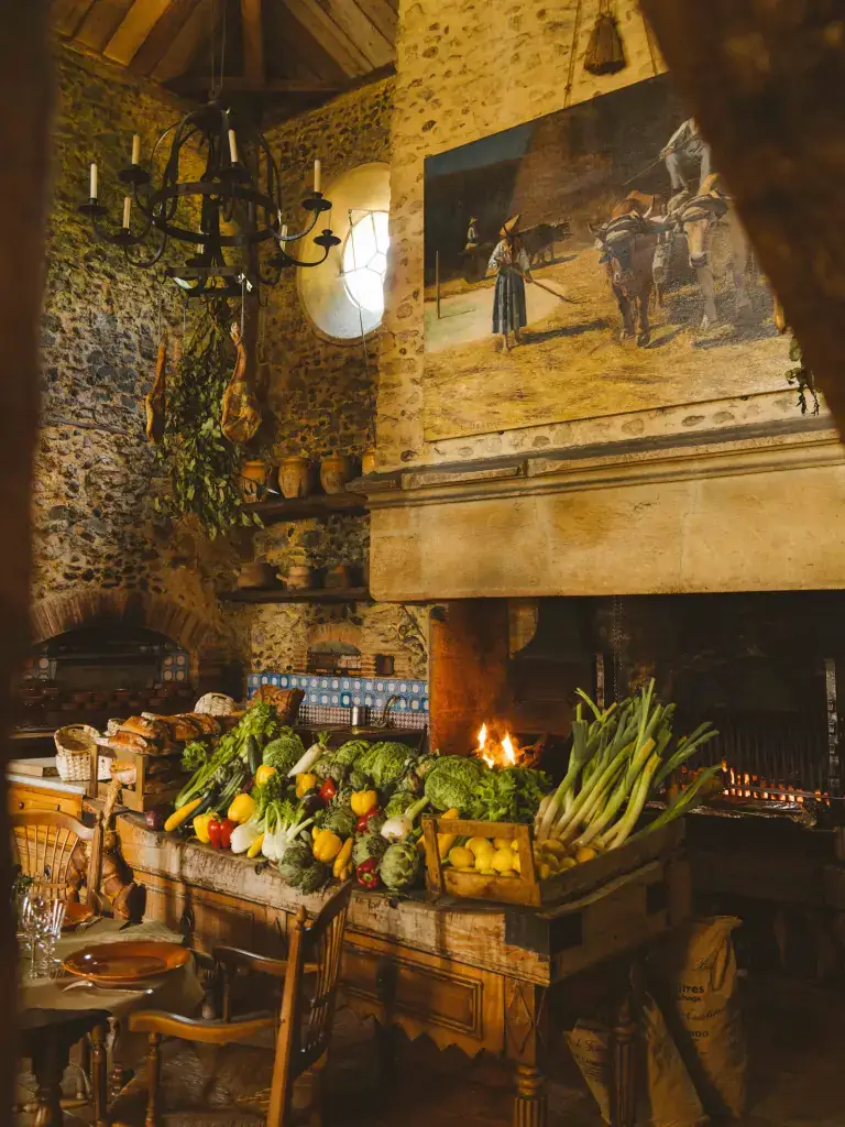 La salle de La Ferme aux Grives aux Prés d'Eugénie, billot couvert de légumes de saison devant la cheminée monumentale où rôtissent les pièces de viande, murs de galets et jambons suspendus.
