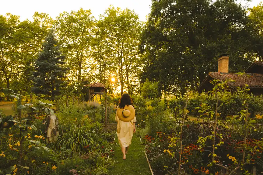 Les jardins potagers des Prés d'Eugénie au soleil couchant, une promeneuse en chapeau de paille sur une allée engazonnée.