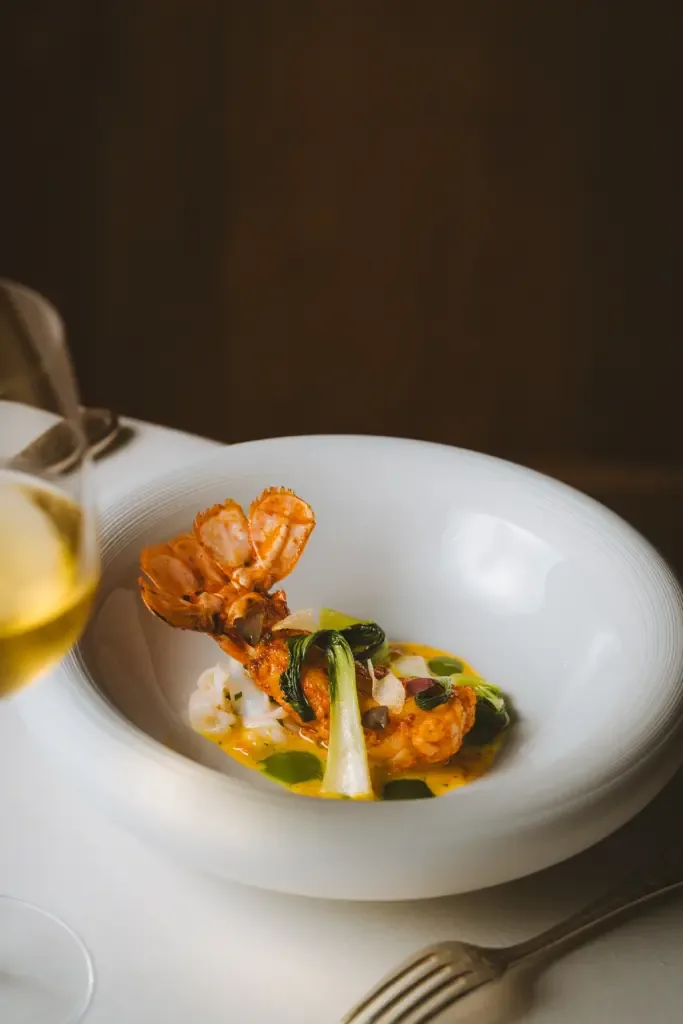 Langoustine dorée au beurre d'épices et pak choï dans un bouillon safrané, assiette blanche et verre de vin blanc, au restaurant Michel Guérard des Prés d'Eugénie.